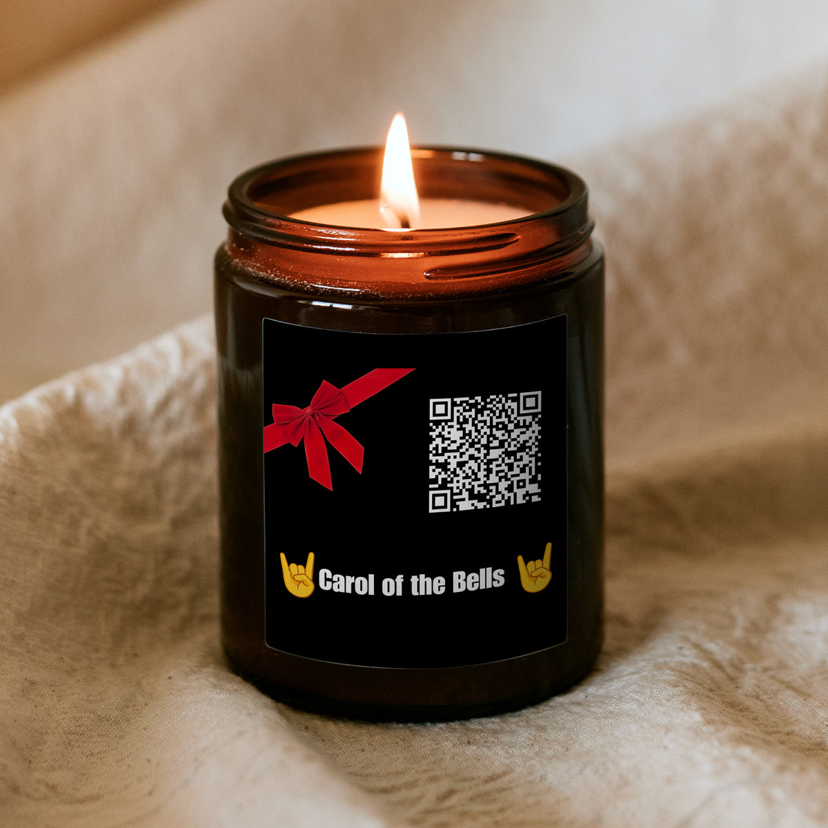 Hořící vánoční svíčka „Melodická“ Kiskedves v hnědé sklenici na měkké dece, na černé etiketě červená mašle, QR kód a nápis Carol of the Bells s rockovými gesty