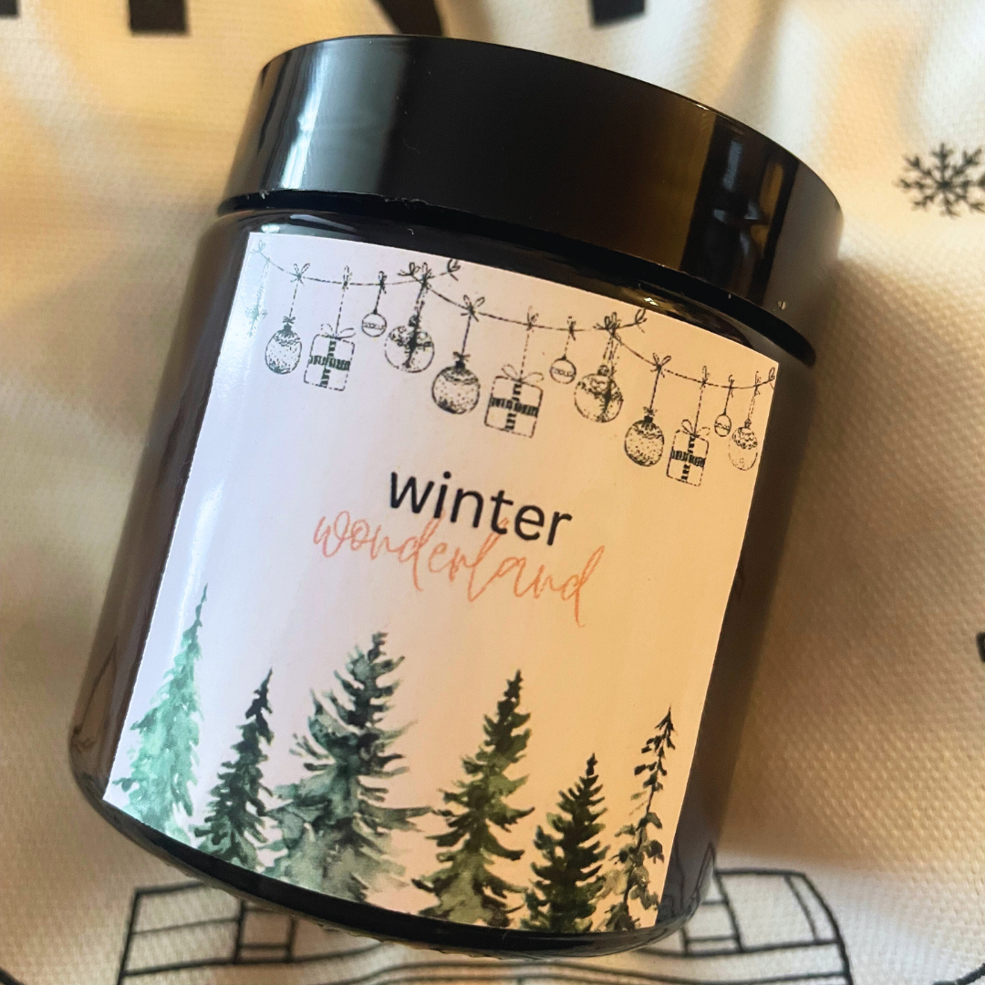 Svíčka Kiskedves Winter Wonderland na textilii s vločkami, zblízka se štítkem s lesem.