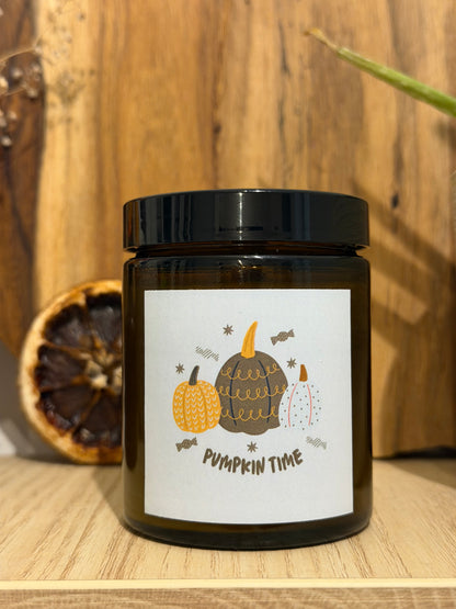 Zavřená sójová svíčka Pumpkin Spice v hnědé sklenici s černým víčkem, se štítkem „Pumpkin time“ s kreslenými dýněmi, na dřevěné polici, vedle ní sušené kolečko pomeranče