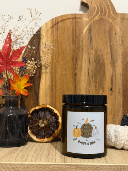 Sójová svíčka Pumpkin Spice v hnědé sklenici se štítkem „Pumpkin time“, před dřevěným prkénkem ve tvaru dýně, vedle váza se sušenými květinami, podzimní listí a bílá háčkovaná dýně
