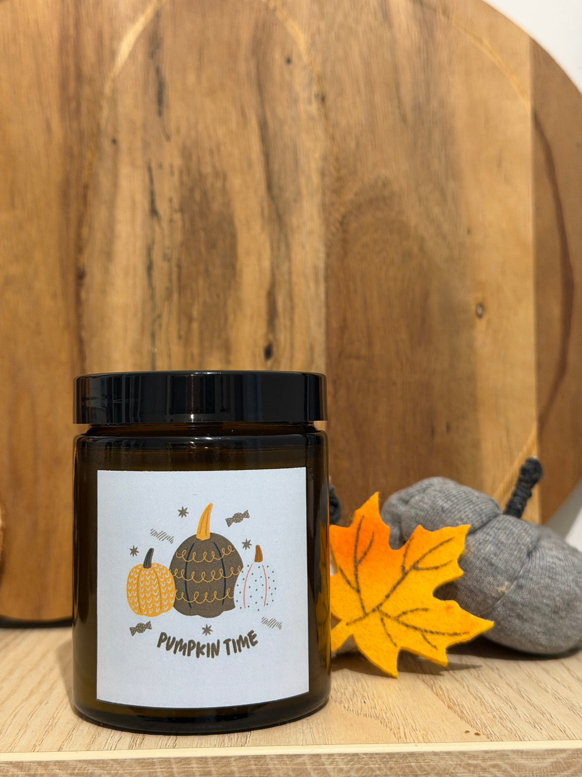 Sójová svíčka Pumpkin Spice v hnědé sklenici se štítkem „Pumpkin time“, před kulatým dřevěným prkénkem, vedle žlutý plstěný javorový list a šedé látkové dýně