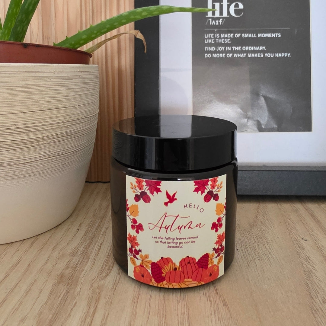 Zavřená sójová svíčka Pumpkin Spice v hnědé sklenici s černým víčkem, se štítkem „Hello Autumn“ s podzimním květinovým věncem, na dřevěné polici, vedle aloe vera v květináči a zarámovaný obrázek