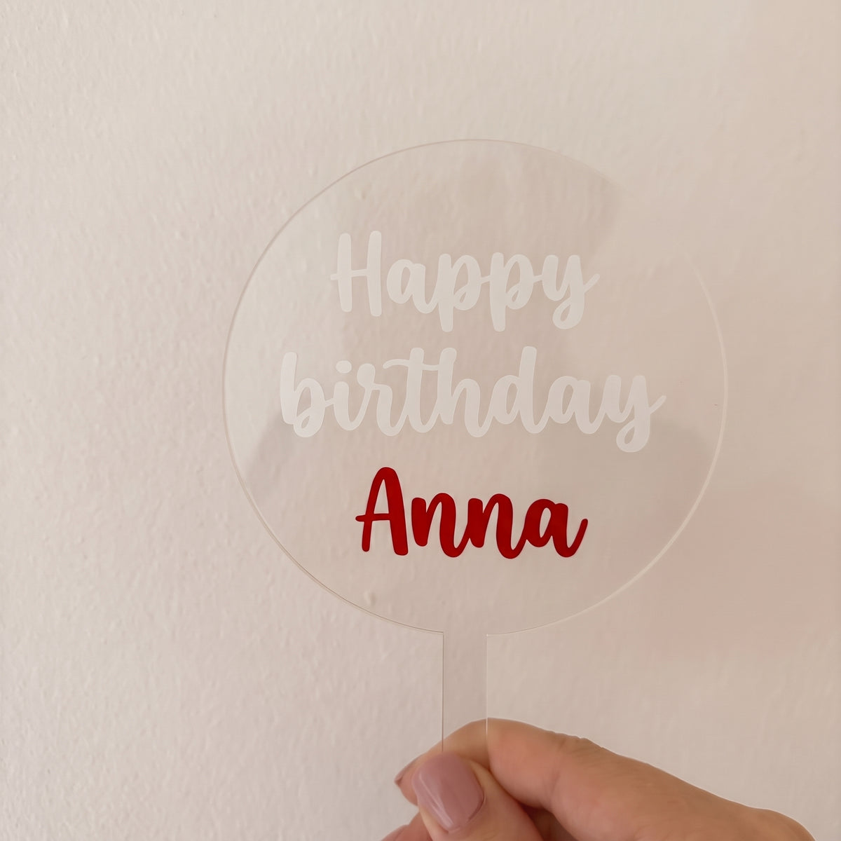Průhledný akrylový zápich na dort s bílým nápisem „Happy birthday“ a červeným jménem „Anna“ v ruce