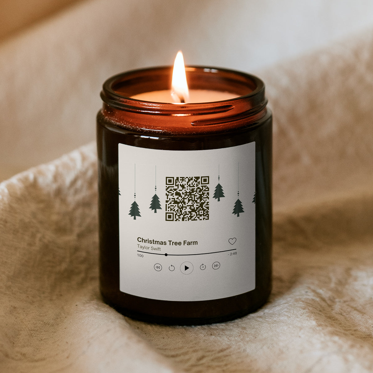 Hořící vánoční svíčka „Melodická“ Kiskedves v hnědé sklenici na měkké dece, na etiketě QR kód a hudební karta s písní Christmas Tree Farm