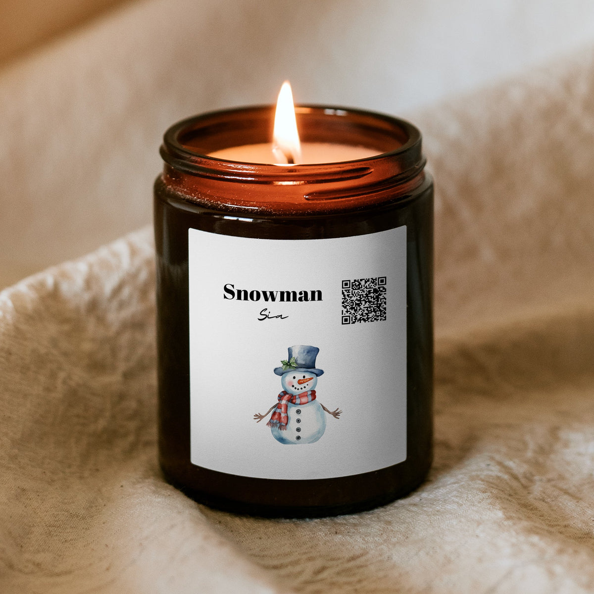 Hořící vánoční svíčka „Melodická“ Kiskedves v hnědé sklenici na měkké dece, na etiketě QR kód, nápis Snowman a akvarelový sněhulák